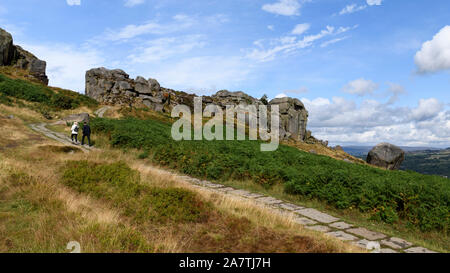 Menschen zu Fuß auf dem Weg zu hohen Sonnenbeschienenen Felsen unter blauen Himmel - malerische Landschaft von Kuh und Kalb Felsen, Ilkley Moor, West Yorkshire, England, UK. Stockfoto