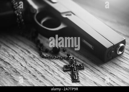 Ein schwarz-weißes Bild einer Waffe, die auf einer hölzernen Oberfläche entlang mit einem schwarzen Katholischen Kreuz liegt, mit Krieg und Religion. Stockfoto