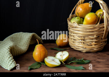 Einen Weidenkorb mit reife Birnen steht auf einem Holztisch. In der Nähe befinden sich drei Birnen. Eine Birne halbiert. Im Hintergrund ist eine grüne Serviette. Stockfoto