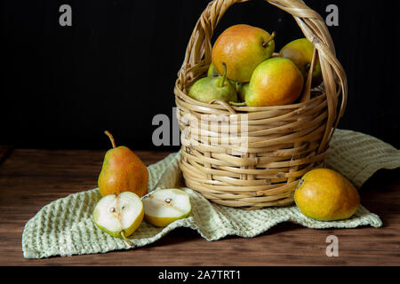 Einen Weidenkorb mit reife Birnen steht auf einem Holztisch. In der Nähe befinden sich drei Birnen. Eine Birne halbiert. Unter dem Korb ist ein grünes Handtuch. Stockfoto