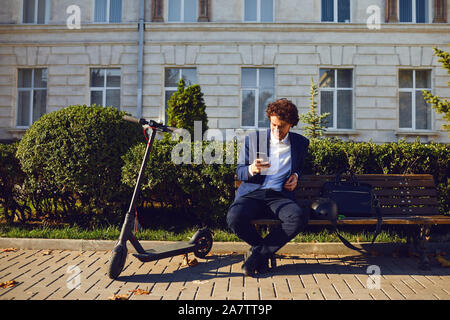 Ein junger curly Mann sitzt auf einer Bank mit einem elektrischen scooter Stockfoto