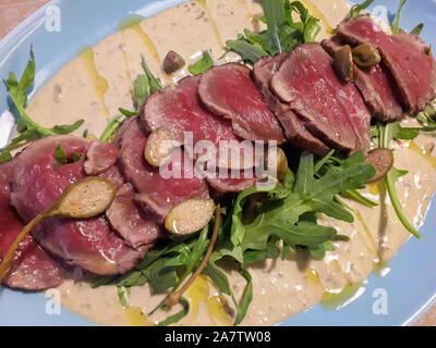 Dünne Scheiben geschnitten mariniertes Rindfleisch mit Thunfisch Sauce serviert, grün Rucola Blätter und Olivenöl auf blauen Platte. Traditionelle italienische Gericht namens Vitello Tonnato. Stockfoto