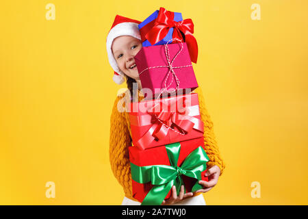 Happy kleines Mädchen hält eine Menge Boxen mit Weihnachten Geschenke. Baby in santa claus Hut auf gelben Hintergrund. Stockfoto