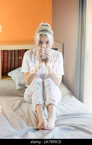 Junge hübsche blonde Frau sitzt auf dem Bett mit einer Tasse Kaffee tragen Schlafanzug in häuslicher Atmosphäre Stockfoto