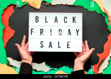 Lightbox Text 'Black Friday Sale" in verbranntem Papier Loch in Weiß, Rot, Gelb und Grün Papier mit verbrannte Kanten, schwarzes Papier kreative Hintergrund, Hände Stockfoto