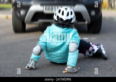 Ein kleines Mädchen in eine blaue Jacke und Helm auf Rollen fiel vor ein fahrendes Auto auf einer Straße der Stadt Stockfoto