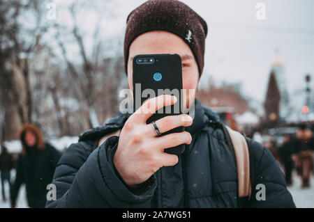 Closeup Bild einer jungen Kerl mit einem Smartphone während des Tages auf ein Winter Straße. Der Kerl ist das Aufnehmen von Fotos. Konzepte der sozialen Netzwerke Stockfoto