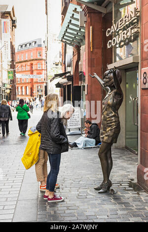 Touristen prüfen ein Cilla Black Statue im Cavern Quarter von Liverpool, Großbritannien, mit einem Bettler im Hintergrund. Stockfoto