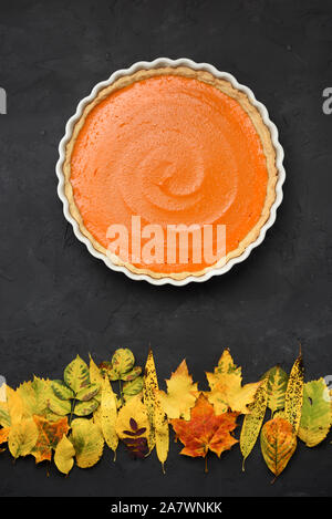 Helles Orange Pumpkin Pie und trockene Blätter wie Sonnenuntergang über Herbst Wald auf schwarzem Hintergrund. Gesunde natürliche Süßigkeiten Konzept, Ansicht von oben Stockfoto