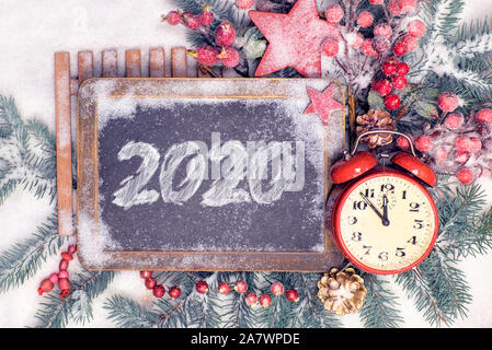 Neues Jahr flach mit 2020 auf der Tafel. Rot und Grün traditionelle winter Dekorationen, Schmuck, Stern, Beeren und Pine Cone auf Schnee. Wecker sh Stockfoto