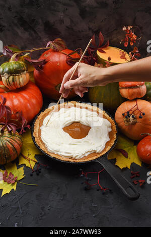 Thanksgiving Konzept. Schlanke Frau Hand pumpking verzieren hausgemachte Kuchen mit Schlagsahne mit vielen Kürbisse und Blätter im Herbst serviert auf Schwarz zurück Stockfoto