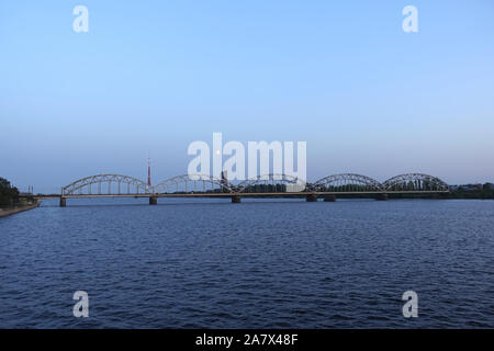 Lange metal Railroad gewölbte Brücke über den Fluss Daugava in Riga gegen allein Gebäude, Fernsehturm und hellen Mond, Blick auf Sonnenuntergang Himmel stand Stockfoto