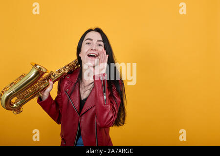 Junge Frau auf der Suche Kamera schreien mit den Händen in der Nähe von Mund hält Saxophon Stockfoto