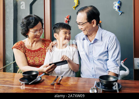 Happy vietnamesischen Jungen zeigen Spiel der Bildschirm des Smartphones zu Großeltern, wenn Sie sitzen im Cafe Tabelle Stockfoto