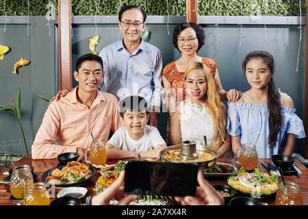 Gerne grosse vietnamesische Familie am grossen Tisch für Foto auf dem Smartphone posing Stockfoto