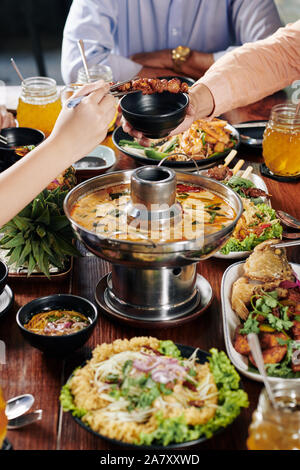 Hände von Menschen und köstliches asiatisches Essen wie squid Suppe, Fisch, gebratene Garnelen und Spieße zum Abendessen Stockfoto