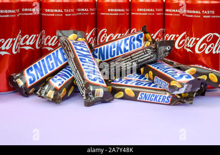 KHARKOV, UKRAINE - OKTOBER 2, 2019: Snickers Schokoriegel in Braun Verpackung liegt auf helle violette Hintergrund mit Coca Cola Dosen hautnah. Famou Stockfoto