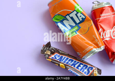 KHARKOV, UKRAINE - OKTOBER 2, 2019: Snickers Schokoriegel verpacken liegt mit leeren roten Coca Cola und Orange Fanta Dosen auf lila Pastell Hintergrund Stockfoto