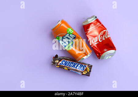 KHARKOV, UKRAINE - OKTOBER 2, 2019: Snickers Schokoriegel verpacken liegt mit leeren roten Coca Cola und Orange Fanta Dosen auf lila Pastell Hintergrund Stockfoto