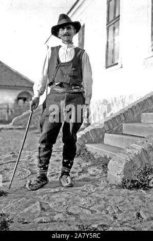 Serbische Mann mit Hut, Hemd, Hose, Gürtel und spitzen Schuhe mit Zuckerrohr stehen außerhalb der Gebäude in Jugoslawien c 1918-1924 Stockfoto