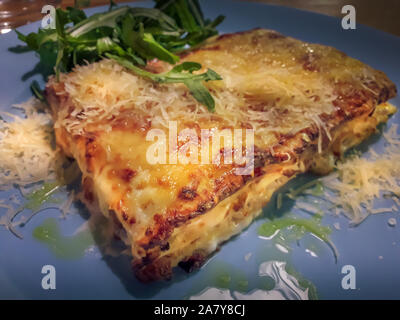 Großer Teil der traditionelle italienische Lasagne durch die Kruste des geschmolzenen Parmesan Käse bedeckt. Es ist auf die blaue Platte mit grünen Rucola Salat serviert, g Stockfoto