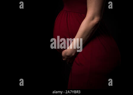 Schwangere Frau im roten Kleid Bauch Holding auf schwarzem Hintergrund. Portrait von aufgeregt, fröhlich, blond, junge Frau in der Schwangerschaft studio schießen. Konzept der mat Stockfoto