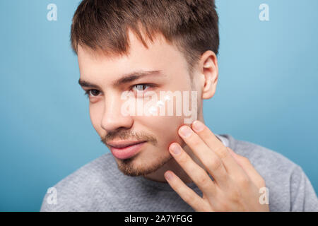 Porträt der jungen brünetten Mann mit dem kleinen Bart und Schnurrbart Anwendung Gesichtscreme unter Augen, Behandlung oder Feuchtigkeitscreme Kosmetik, Hautpflege Konzept. Ich Stockfoto