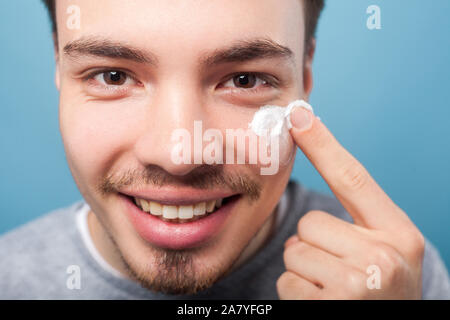 Männliche Hautpflege Kosmetik. Portrait von glücklich aussehenden Mann mit dem kleinen Bart und Schnurrbart Anwendung Creme oder anti-aging Kosmetik auf Gesicht, auf der Suche nach Kameras Stockfoto