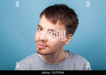 Hautpflege Routine. Portrait von fröhlichen brünett Mann mit dem kleinen Bart lächelnd an Kamera, in Flecken von Feuchtigkeits-creme unter Augen, männliche Gesichtszüge cosm Stockfoto