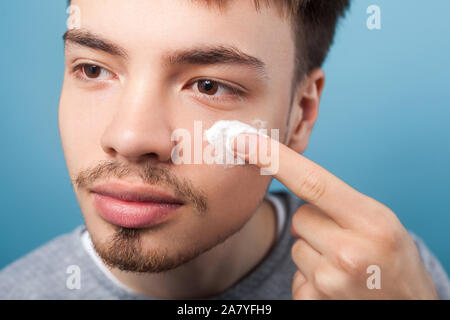 Männliche Schönheit und Hautpflege. Porträt junger stattlicher Mann mit kleinem Bart und Schnurrbart Anwendung Creme oder anti-aging Kosmetik auf Gesicht, beiseite. Stockfoto