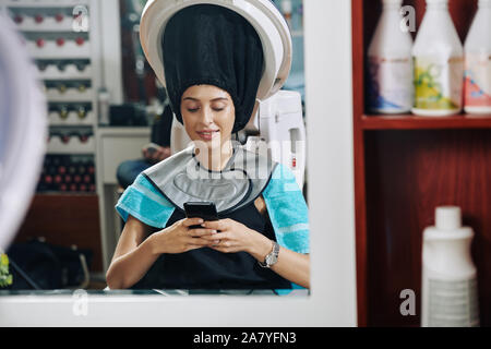 Lächelnde junge Frau liest Textnachrichten von ihren Freunden, wenn sie unter Haar Dampfgarer in Beaty Salon sitzen Stockfoto