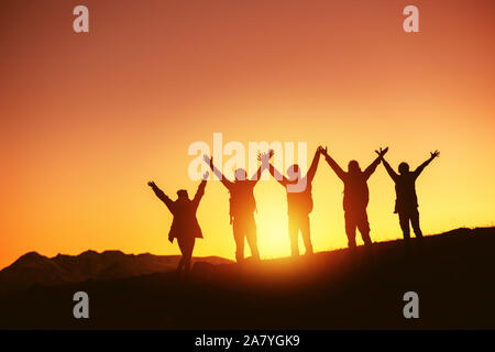 Gruppe der glücklichen Völker Silhouetten steht mit erhobenen Armen gegen Sonnenuntergang und Berge Stockfoto