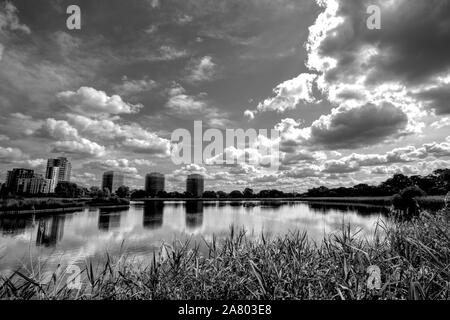 Woodberry Feuchtgebiete, Stoke Newington, London. See mit Gras im Vordergrund, die Gebäude im Hintergrund, Moody Wolken im Himmel und Reflexionen. Stockfoto