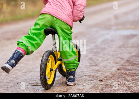Ansicht der Rückseite des unteren Bereich eines Kindes Mädchen erhalten auf ihre Balance Fahrrad in warme und wasserdichte Kleidung auf einem feuchten und verschmutzten Schotterstraße im Herbst. Gesehen Stockfoto