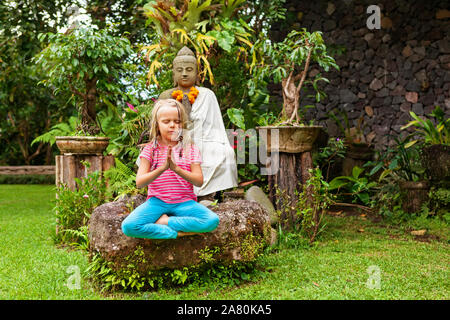 Morgen lustig Meditation in den tropischen Garten. Glückliches Mädchen sitzen, im Yoga Lotus entspannen Pose mit Mudra namaste Hände. Aktives Kind gesunde Lebensweise. Stockfoto