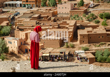 Arabische Frau in Rot sieht auf das Mobil in der Kasbah Ait Ben Haddou gekleidet in Ouarzazate, Marokko Oktober 2019 Stockfoto