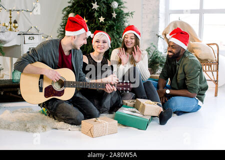 Bild zeigt eine Gruppe von vier Freunden Weihnachten zu Hause feiern. Jungen kaukasischen Mann ist, Gitarre zu spielen und die Mädchen und afrikanischer Mann lächeln Stockfoto