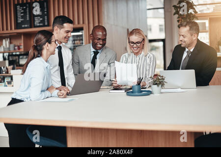 Lächelnd Gruppe verschiedener Geschäftsleute mit Laptops und über Schreibarbeit beim Stehen um einen Tisch während einer Sitzung in einem modernen Büro Stockfoto