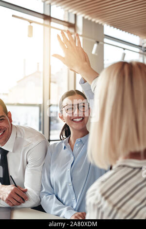 Zwei junge Unternehmerinnen lachen und hohe FIVING zusammen bei einem Treffen mit einem männlichen Kollegen an einem Tisch in einem modernen Büro Stockfoto