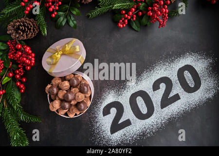 Wir begrüßen das Jahr 2020 mit einer Schachtel Pralinen und Schokolade Trüffel, einige Ananas und einige holly Filialen auf einem dunklen Hintergrund. Stockfoto