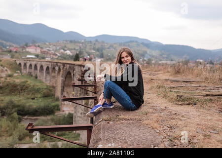Junge touristische Mädchen sitzen auf Viadukt mit alten Eisenbahnschienen. Alte Eisenbahnviadukt im Mountain Resort Dorf Vorokhta. Die Ukraine, die Karpaten. Stockfoto