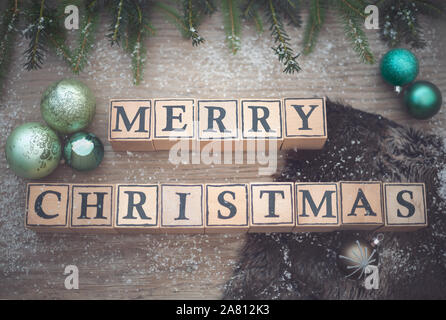Holzwürfel mit Buchstaben Form der englische Text frohe Weihnachten. Sie liegen auf einem Holz Hintergrund. Tannenzweigen und Weihnachten Kugeln sind. Stockfoto