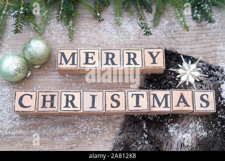 Holzwürfel mit Buchstaben Form der englische Text frohe Weihnachten. Sie liegen auf einem Holz Hintergrund. Tannenzweigen und Weihnachten Kugeln sind. Stockfoto