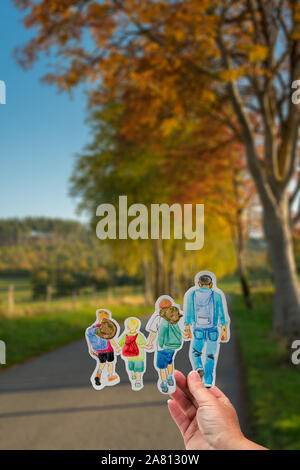 Eine gepflasterte Straße führt durch sonnige Herbst Landschaft mit orange verlässt. Hand hält Zeichnung mit Wandern Familie mit Rucksäcken. Stockfoto