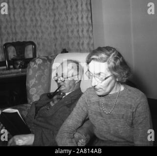 1950er Jahre, historisch, in einem vorderen Zimmer, ein älterer Mann in Anzug und Krawatte, der in einem Sessel sitzt und Pfeife raucht und ein Buch liest, während seine Frau neben ihm sitzt. Stockfoto