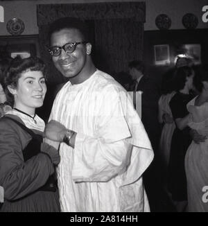 1950er Jahre, historisches, Nachkriegs-Großbritannien und eine junge englische Dame, die mit einem Überseemänner aus Westafrika tanzt, der ein traditionelles, lang fließendes Gewand trägt, eine Agbada, die normalerweise von Männern in Nigeria und anderen westafrikanischen Ländern, London, England, Großbritannien getragen wird. In dieser Ära kamen viele Völker aus den überseeischen Commonwealth-Ländern nach Großbritannien, um zu arbeiten und zu studieren und Funktionen und Tänze zu spielen und wurden gehalten, um sie in die britische Lebensweise einzuführen. Stockfoto