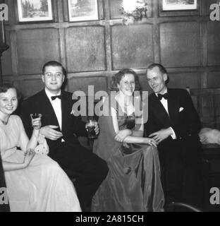 1950er Jahre, historisch, zwei glückliche Paare, die an einer formellen Veranstaltung mit schwarzer Krawatte teilnehmen, sitzen bei einem Drink in einem holzgetäfelten Veranstaltungsraum in England, Großbritannien. Stockfoto