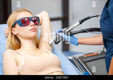 Kosmetologie Service. Junge womanin Schutzbrille unter Beauty Klinik lag, blickte nachdenklich, während der Arzt mit Laser Entfernen der Haare auf achselhöhle schließen Stockfoto