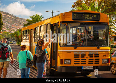 Menschen, die an einem sonnigen Tag mit Palmen und Blick auf die Hügel in Funchal, Madeira, Portugal, in einen gelben öffentlichen Bus steigen Stockfoto