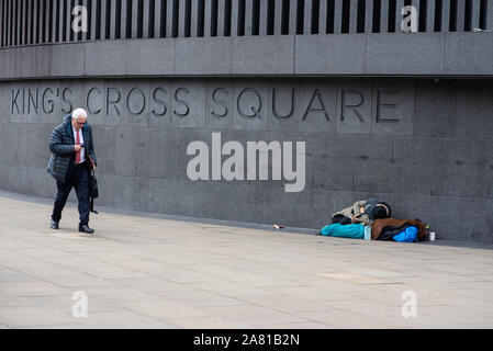 London, Großbritannien - 5 November, 2019: ein Mann im Anzug Spaziergänge durch King's Cross entfernt, während eine grobe Sleeper auf dem Bürgersteig liegt. Stockfoto